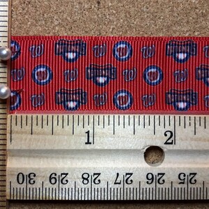 May include: Red and white ribbon with a repeating pattern of a blue and white baseball logo. The ribbon is measured against a wooden ruler, showing it is approximately 2 inches long.