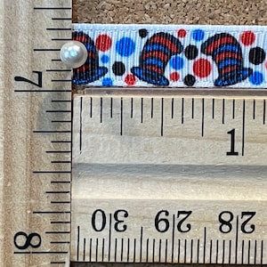 May include: White ribbon with a repeating pattern of red, blue, and black hats and polka dots. The ribbon is measured with a wooden ruler, showing inches. A pearl pin is used to hold the ribbon in place.