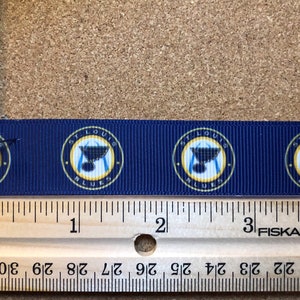 Pode incluir: Um close-up de uma fita azul com um padr&atilde;o repetido do logotipo do St. Louis Blues. O logotipo apresenta um c&iacute;rculo dourado com as palavras "St. Louis Blues" e um taco de h&oacute;quei com um trof&eacute;u da Stanley Cup no topo. A fita &eacute; medida com uma r&eacute;gua, mostrando que tem 10 cm de comprimento.