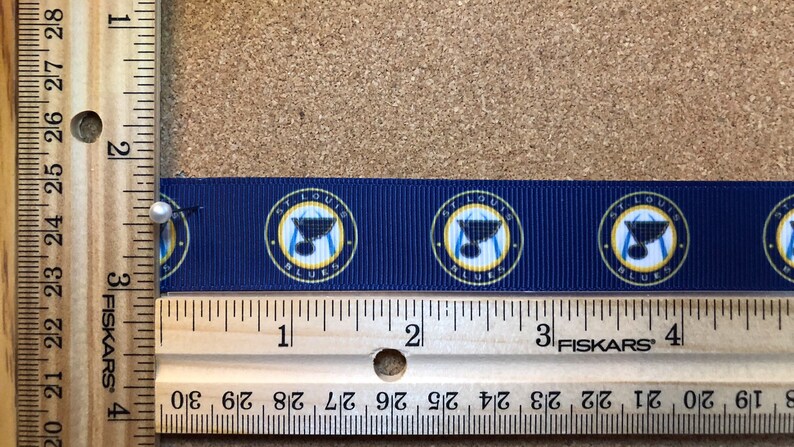 Pode incluir: Um close-up de uma fita azul com um padr&atilde;o repetido do logotipo do St. Louis Blues. O logotipo apresenta um c&iacute;rculo dourado com as palavras "St. Louis Blues" e um taco de h&oacute;quei com um trof&eacute;u da Stanley Cup no topo. A fita &eacute; medida com uma r&eacute;gua, mostrando que tem 10 cm de comprimento.