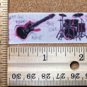 Puede incluir: Cinta blanca con un diseño de guitarra, batería y altavoz negro y rosa. La cinta se mide con una regla, mostrando que tiene aproximadamente 2,5 cm de ancho.