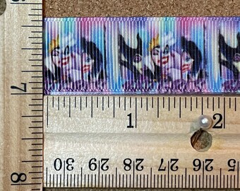 Pode incluir: Fita estampada com um padrão repetido de um personagem de desenho animado em tons de roxo, azul e branco. A fita é medida com uma régua de madeira, mostrando marcações em centímetros.