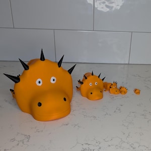 以下が含まれることがあります： 黒いトゲと白い目を持つ、オレンジ色の漫画のような生き物の彫刻。彫刻のサイズは異なり、最大のものが前景にあります。背景には白いタイルの壁と白いカウンタートップがあります。