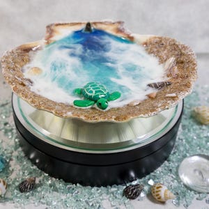 May include: A decorative seashell with a miniature ocean scene. The shell features a blue and white wave design with a green turtle figurine. Small seashells and blue glass beads surround the base.
