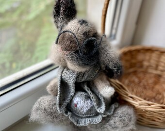 May include: A gray knitted bunny wearing glasses and a gray knitted dress. The bunny is sitting on a windowsill with a wicker basket in the background.