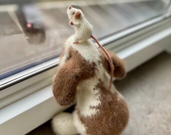 May include: A brown and white knitted dog toy with a white knitted mouse on top. The dog toy is standing on a white windowsill.