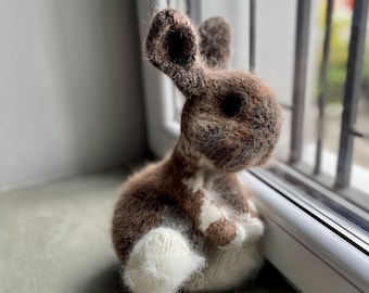 May include: A handmade, brown and white knitted bunny rabbit figurine. The bunny is sitting near a window, with its ears perked up. The bunny has a fluffy white tail and is made of soft yarn.