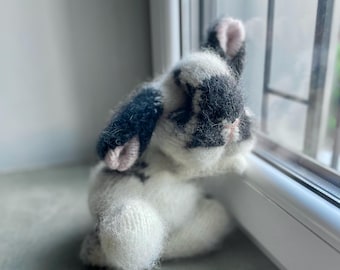 May include: A handmade, plush bunny rabbit in a sitting position, with a black and white speckled pattern. The bunny has pink inner ears and is positioned near a window, creating a cozy, soft toy aesthetic.