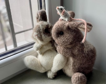 May include: A pair of knitted bunny toys, one cream and the other brown and white, with a small grey mouse perched on top. The toys are handmade, with a soft, textured appearance. The scene is set near a window, giving a cosy feel.