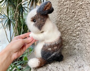 May include: A hand-knitted plush toy rabbit with brown, white, and gray fur. The rabbit is sitting on a gray textured surface.