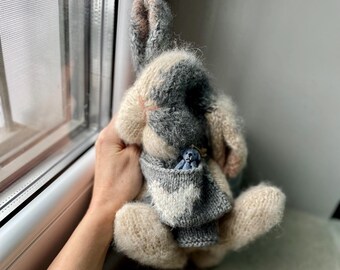 May include: A gray and white knitted bunny rabbit toy with a smaller blue knitted bunny toy in its pocket. The larger bunny is holding its arms up and has a long, floppy ear.