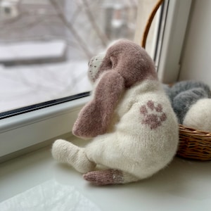 May include: A knitted plush dog toy with a brown head and white body. The dog has a brown paw print on its back. The toy is sitting on a white surface near a window.