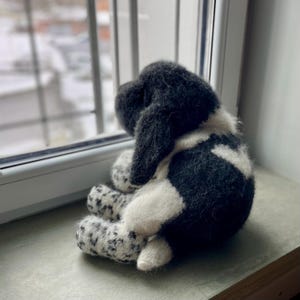 May include: A handmade plush dog toy in black and white, sitting on a windowsill. The dog is facing away from the camera, looking out the window. The toy has a soft, textured appearance.