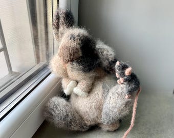 May include: A handmade, knitted cat toy in shades of beige, brown, and black, with white paws, is positioned near a window. A small, gray knitted mouse with pink ears and a long pink tail sits nearby. The cat appears to be looking out the window.
