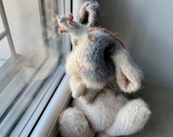 May include: A knitted gray and white bunny rabbit with pink ears and a long tail. The bunny is sitting on a windowsill and looking out the window.