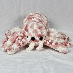 May include: A crocheted plush toy shaped like a spider with pink and white yarn. The spider has large black eyes and white crocheted fangs.
