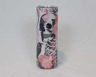 May include: A white tumbler with a black and white skull and skeleton design with pink floral accents. The skull has a crown of pink flowers and a black bird is perched on the skeleton's shoulder.