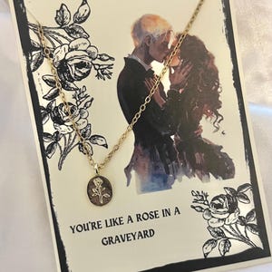 K&ouml;nnte beinhalten: Eine goldfarbene Halskette mit einem kleinen ovalen Anh&auml;nger mit einem Rosendesign. Die Halskette wird auf einer Karte mit schwarzem Rand, einer romantischen Illustration und dem Text "YOU'RE LIKE A ROSE IN A GRAVEYARD" pr&auml;sentiert.