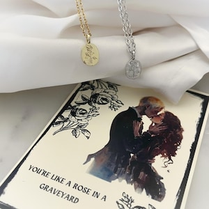 K&ouml;nnte beinhalten: Zwei Halsketten mit Rosenanh&auml;ngern, eine goldfarbene und eine silberfarbene, liegen auf wei&szlig;em Stoff. Darunter eine Karte mit einer romantischen Illustration eines sich k&uuml;ssenden Paares und dem Text "YOU'RE LIKE A ROSE IN A GRAVEYARD."