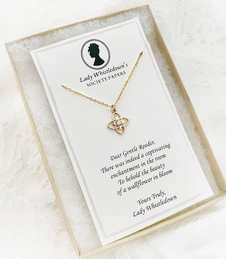 May include: A gold necklace with a delicate, ornate pendant featuring a clear crystal in the center. The necklace is displayed in a white box with a card that reads "Lady Whistledown's Society Papers" and a quote about a wallflower in bloom.