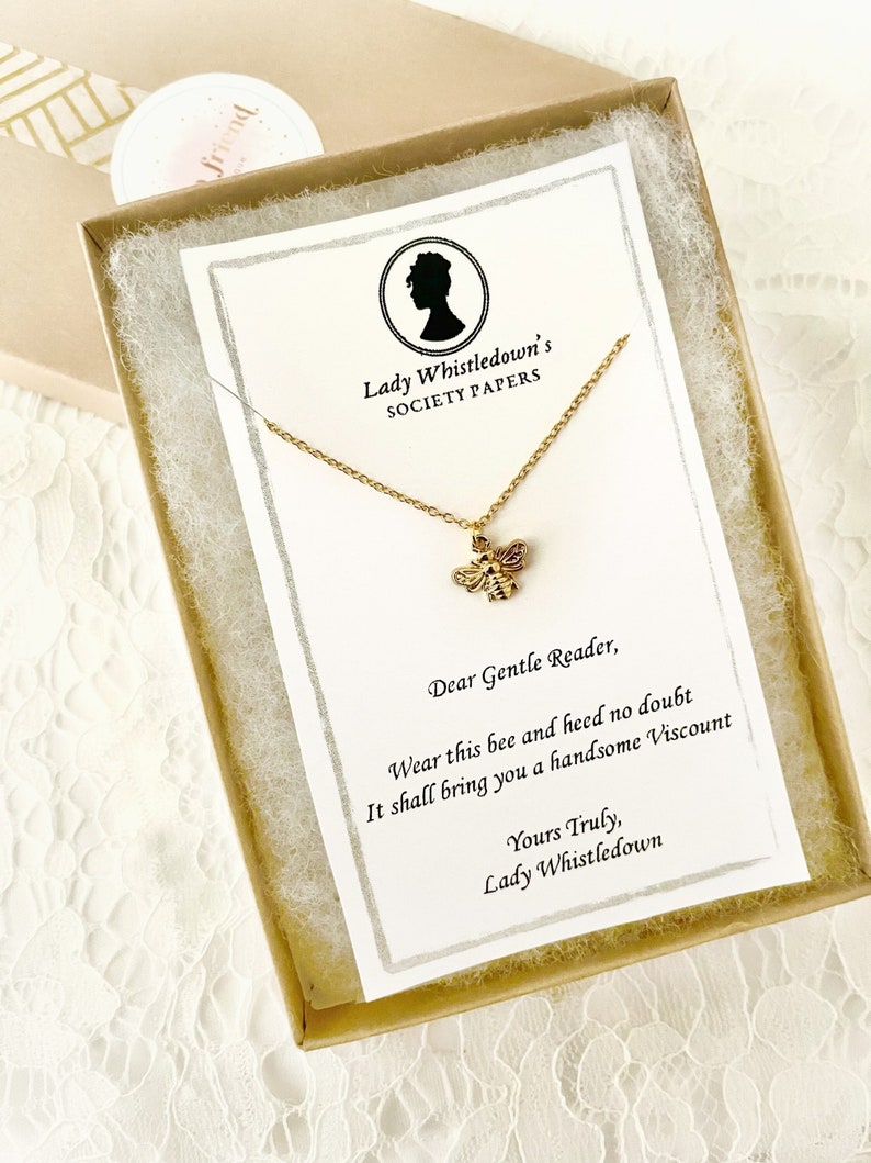 May include: A gold necklace with a bee charm in a small box. The box has a card inside with the text "Lady Whistledown's Society Papers" and a message that reads "Dear Gentle Reader, Wear this bee and heed no doubt It shall bring you a handsome Viscount Yours Truly, Lady Whistledown".
