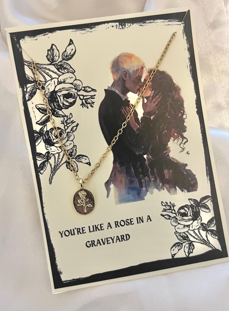 K&ouml;nnte beinhalten: Eine goldfarbene Halskette mit einem kleinen ovalen Anh&auml;nger mit einem Rosendesign. Die Halskette wird auf einer Karte mit schwarzem Rand, einer romantischen Illustration und dem Text "YOU'RE LIKE A ROSE IN A GRAVEYARD" pr&auml;sentiert.