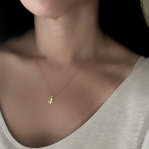 K&ouml;nnte beinhalten: Eine zarte goldene Halskette mit einem kleinen, ovalen Anh&auml;nger. Der Anh&auml;nger hat ein detailliertes Design, und die Kette ist fein und elegant. Die Halskette wird am Hals einer Person getragen, vor einem hellen Oberteil.