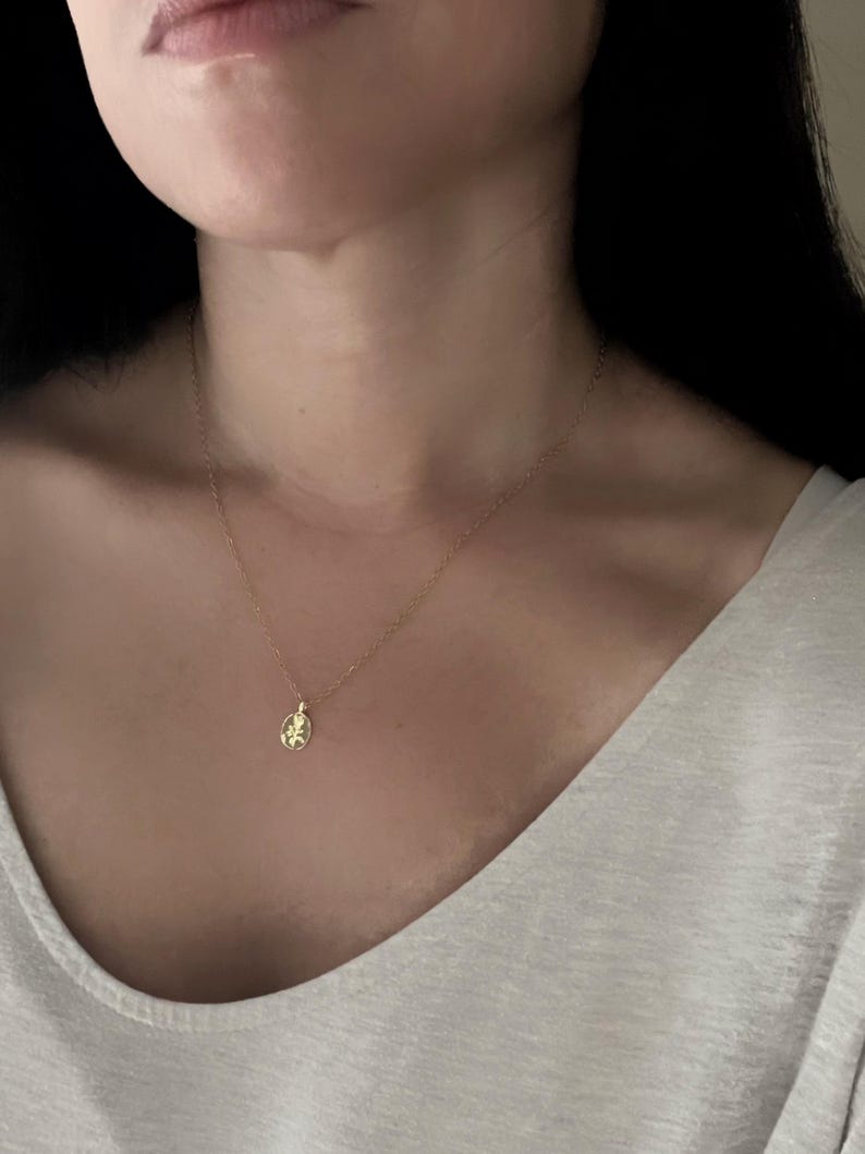 K&ouml;nnte beinhalten: Eine zarte goldene Halskette mit einem kleinen, ovalen Anh&auml;nger. Der Anh&auml;nger hat ein detailliertes Design, und die Kette ist fein und elegant. Die Halskette wird am Hals einer Person getragen, vor einem hellen Oberteil.