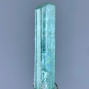 Peut inclure: Un cristal bleu clair translucide avec des stries verticales. Le cristal est de forme prismatique rectangulaire et long, avec un sommet facetté. Il est monté sur un support en métal argenté sur fond gris. Le cristal est légèrement irisé.