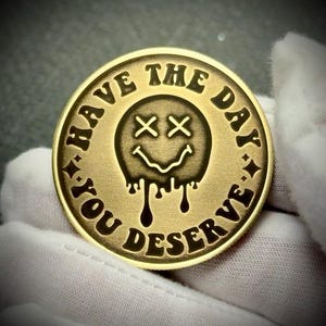 May include: A gold-colored coin with the text "HAVE THE DAY YOU DESERVE" encircling a melting smiley face with crossed-out eyes. The coin is round and has a slightly raised edge, with a dark background.