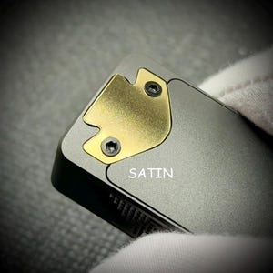 May include: Close-up of a metallic gray object with a gold-colored accent piece secured by two screws. The word "SATIN" is printed in white on the object's surface. The object has a sleek, modern design.