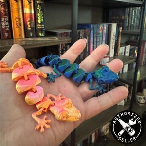 May include: Two 3D printed gecko lizards, one orange and one blue, are held in a person's hand. The lizards are articulated and have a segmented body.