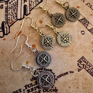 May include: Earrings with compass rose charms in gold and silver tones. The earrings have a hook closure and are displayed on a vintage map background. The compass rose design features directional markers.