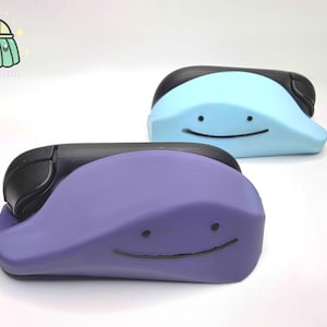 Könnte beinhalten: Zwei amorphe, klecksförmige Figuren mit glatten Oberflächen, eine in Hellblau und eine in Violett. Jede Figur hat einen schwarzen, geschwungenen Mund und zwei kleine schwarze Punkte als Augen sowie ein schwarzes, abgerundetes Teil an der Rückseite.