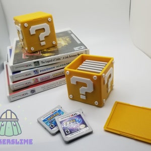 May include: A yellow question mark block with a white question mark and white accents, next to a stack of video game cases. The block is a container for game cartridges. Two game cartridges are in front of the block.