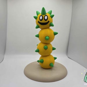Puede incluir: Escultura artesanal de una criatura amarilla apilada con púas verdes. La esfera superior tiene una cara sonriente. La escultura se asienta sobre una base redonda beige. Una creación original y divertida.