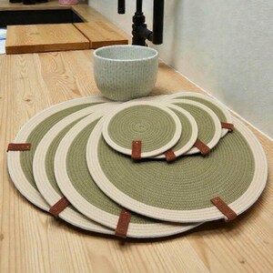 Puede incluir: Un conjunto de manteles individuales y posavasos redondos en tonos verdes y crema. Presentan un diseño de cuerda trenzada con detalles de cuero. El conjunto incluye varias tallas, adecuadas para diversas necesidades de mesa. Los manteles miden unos 30 cm de diámetro.