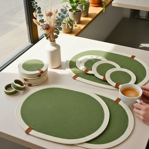May include: A set of green and cream-colored woven placemats, coasters, and napkin rings. The oval placemats and round coasters have a green center with a cream-colored border. Brown leather tags are attached to the placemats. A white vase with dried flowers is also present.