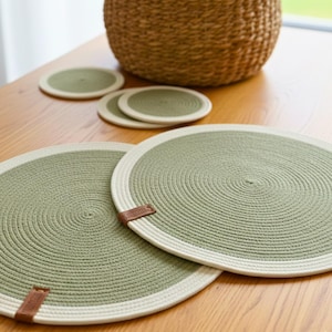 May include: A set of green and white circular placemats and coasters made of woven rope. The placemats have a light green center with a white border and a small brown leather tag. A woven basket is in the background.