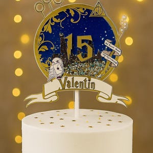 May include: A white cake topped with a custom cake topper. The topper features a blue and gold circular design with the number 15, a silhouette of a castle, and the name "Valentin". Additional elements include a golden snitch and an owl.