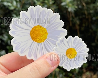 Op de afbeelding: Twee madeliefjesstickers met witte bloemblaadjes en gele centra tegen een groene achtergrond. De stickers hebben de tekst "DESIGNS BY VALAN" erop gedrukt.