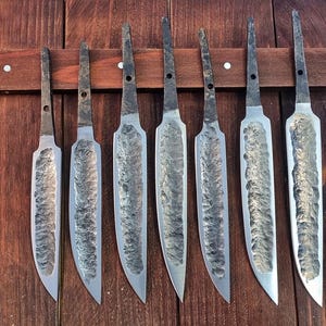 Könnte beinhalten: Sieben handgeschmiedete Messerklingen aus Stahl, die in einer Reihe angeordnet sind. Jede Klinge hat eine dunkle, texturierte, gehämmerte Oberfläche in der Mitte und eine glatte, polierte Schneide. Die Erle sind schmal mit einem Loch.