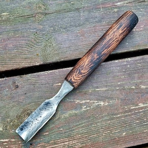 May include: A wood chisel with a dark, stained wooden handle and a silver metal blade. The chisel is angled across a weathered wooden surface, showcasing the tool's design and craftsmanship. The handle's grain is visible.