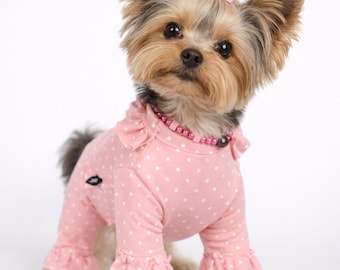 Puede incluir: Un pequeño Yorkshire Terrier lleva puesto un mono de manga larga de color rosa claro con un estampado de lunares blancos. La prenda presenta detalles de volantes en los hombros y en los puños de las mangas. El perro tiene un pequeño lazo rosa con un cristal en el centro sobre la cabeza.