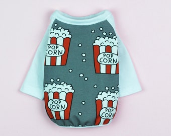 May include: A white dog shirt with a grey body featuring a repeated pattern of red and white popcorn containers with the text "POP CORN" on each container. The shirt has white sleeves and a white collar.