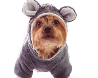 May include: A small brown and tan dog wearing a gray hooded sweater with large ears. The sweater has a white trim around the ears and the hood. The dog is looking directly at the camera.