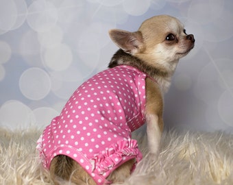 Puede incluir: Un pequeño chihuahua marrón claro con un vestido rosa y blanco de lunares con ribete de volantes. El perro está sentado en una alfombra blanca y esponjosa y mirando hacia la derecha.