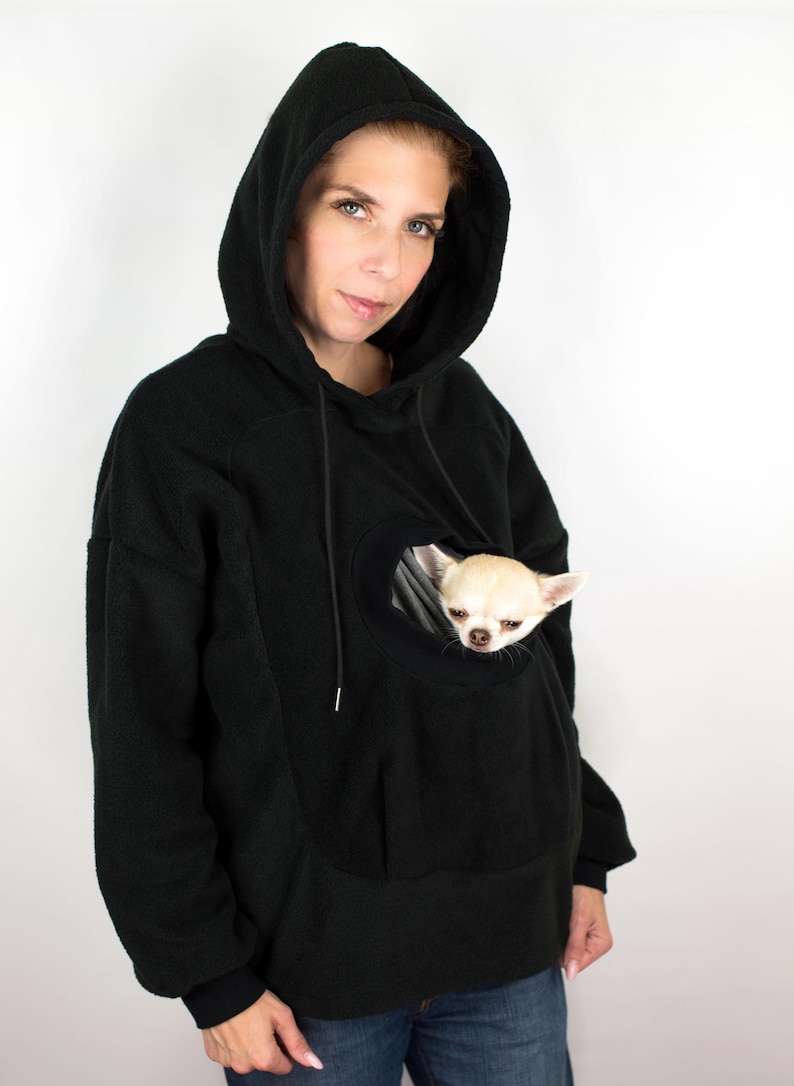 May include: A black fleece hoodie with a large pocket on the front that is designed to hold a small dog. A small white chihuahua is sitting in the pocket.