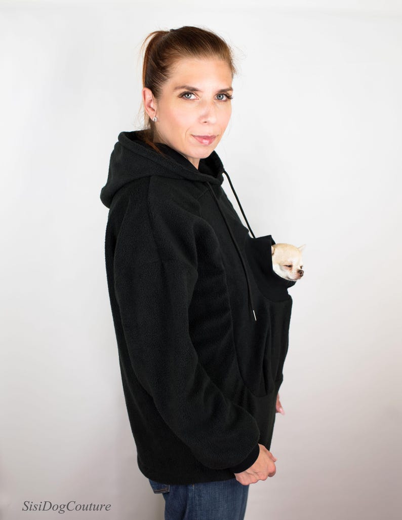 May include: A black fleece hoodie with a large pocket on the front, designed to hold a small dog. A small white dog is peeking out of the pocket.