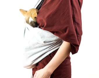 May include: A person wearing a maroon shirt and grey jeans is carrying a small brown and white dog in a grey sling.